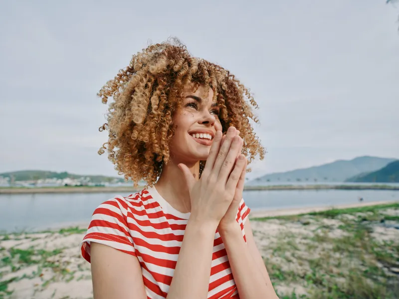 Young woman with curly hair wearing a red striped shirt smiles softly and clasps her hands in prayer by a calm shoreline, enjoying warm sun, blue sky, and gentle breeze.