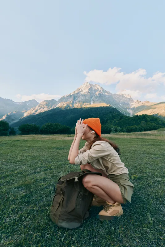 Young woman crouching in a grassy field with a backpack and orange hat, wearing casual wear. A mountain range rises in the background, conveying outdoor lifestyle and travel adventure.