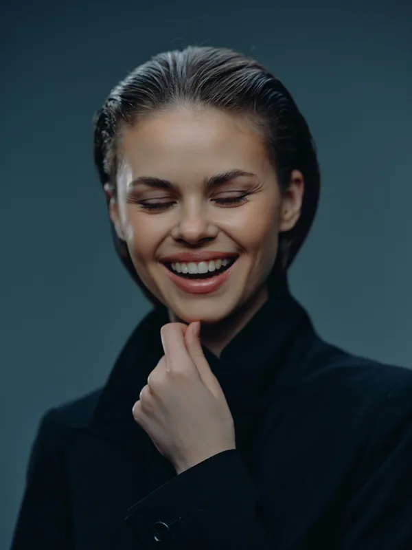 Smiling woman with closed eyes wearing black coat expressing happiness and calmness in dark studio lighting.