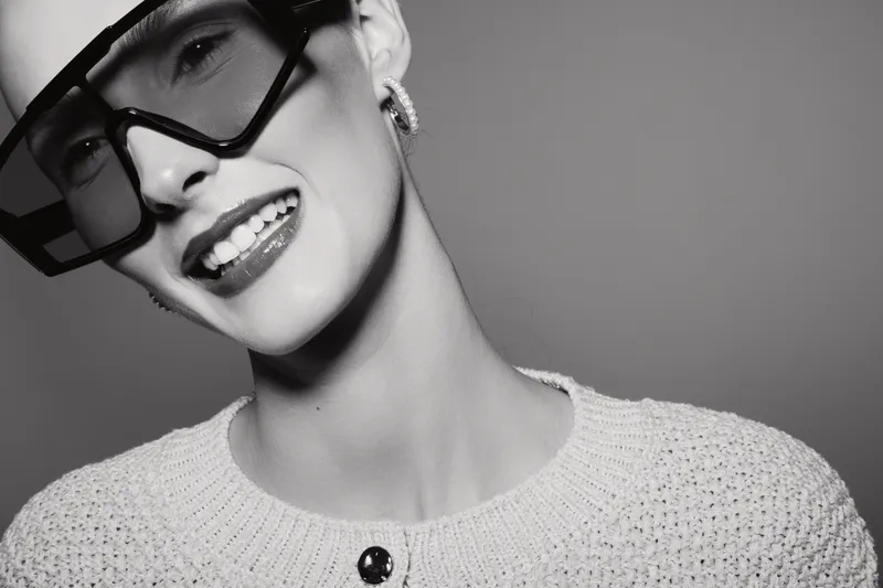 Happy Caucasian woman with light skin wearing oversized sunglasses smiles in a studio. Fashion portrait with monochrome styling and elegant outfit concept.