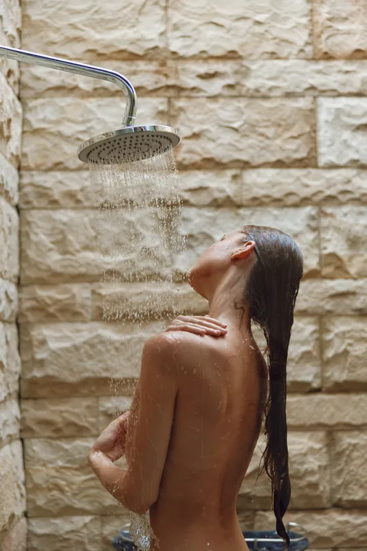 woman showering under rain showerhead with wet hair and relaxed posture, natural light and textured stone wall background in warm tones