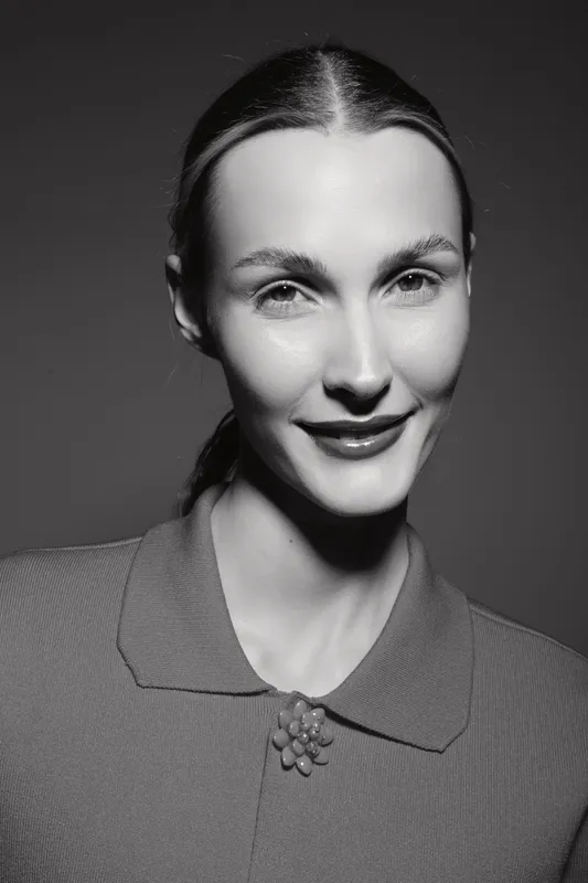 Smiling Caucasian woman with fair skin and slicked back hair poses in textured collar top with brooch. Studio portrait with high contrast lighting and natural expression.