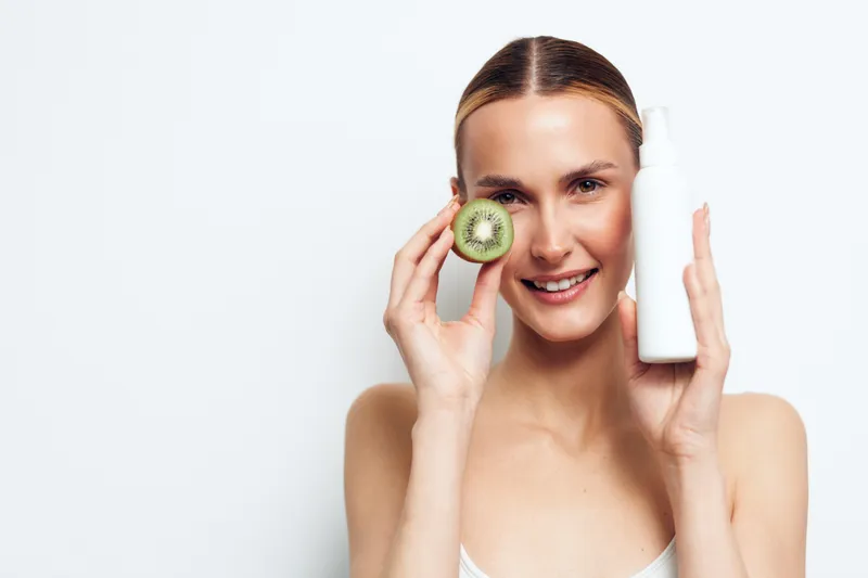 Young woman with clean skin holds kiwi slice near eye and white cosmetic bottle in other hand smiling on white background skincare concept natural beauty and freshness