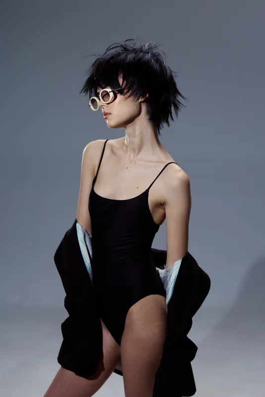 Stylish slim woman in black bodysuit and sunglasses posing with jacket on gray background