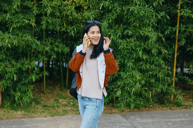 Woman talks on a smartphone while strolling outdoors in casual jacket and jeans, sunny urban vibes, leafy backdrop, candid street style, relaxed mood and contemporary fashion.