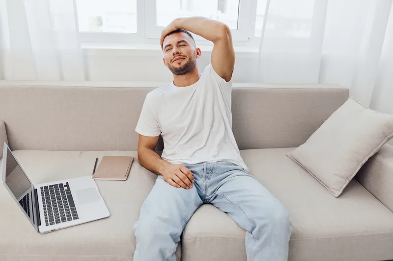 Relaxed young man sitting on the couch, enjoying a moment of leisure with his laptop and a notebook, surrounded by a cozy indoor atmosphere.