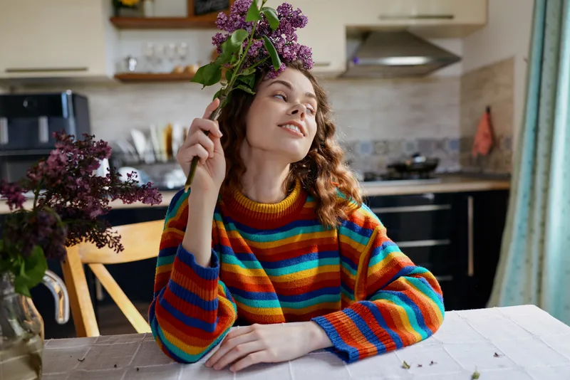 Cheerful woman indoors, sitting at a dining table with a colorful sweater, holding flowers, enjoying a sunny, relaxed moment in a cozy home kitchen, soft natural light and vibrant colors
