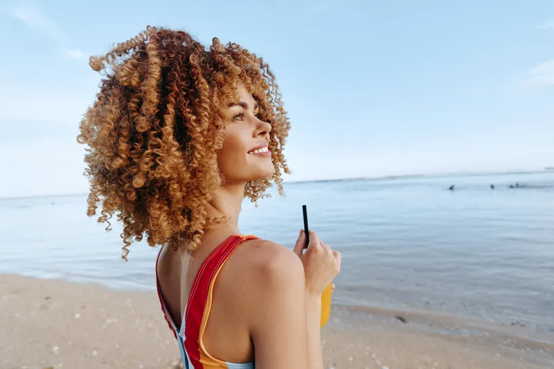 Young woman with curly hair stands on a sunny beach, holding an orange drink and gazing toward the calm sea, wearing a bright red swimsuit and enjoying the breeze.
