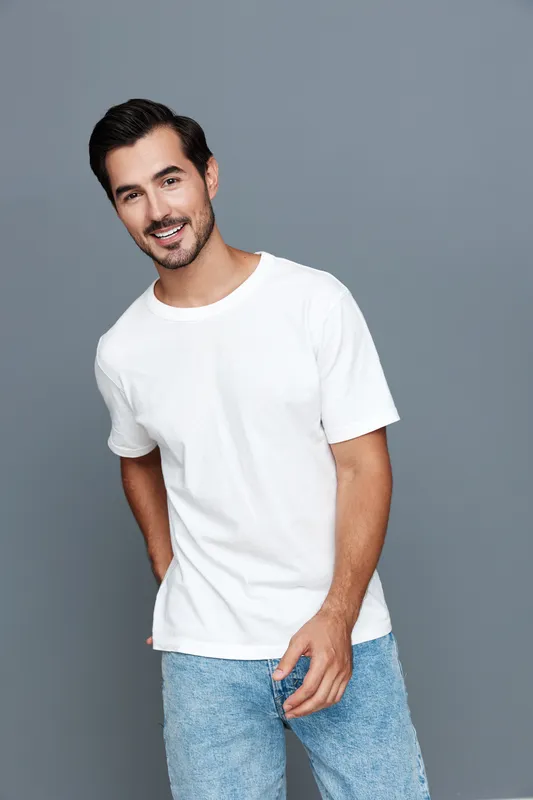 Smiling man in casual white t-shirt poses confidently against a grey background, exuding a cheerful and approachable vibe. Style and personality shine through.