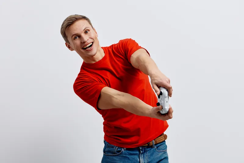 Happy young man with short blonde hair in casual red t-shirt and jeans holding video game controller, expressing excitement and joy, isolated on plain white background. People lifestyle concept
