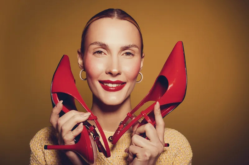 Smiling woman holds red high heels near face, wearing gold earrings and beige sweater. Studio portrait on yellow background. Concept of fashion, style, and confident beauty.