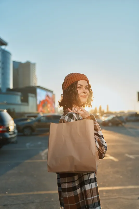 Woman shopping with paper bag in urban parking at sunset, candid lifestyle portrait capturing authenticity, golden hour glow and mindful living with emotional storytelling vibe.