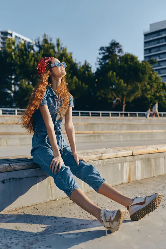 Lifestyle woman in modern boho-western denim street style with red bandana and sunglasses relaxing outdoors under bright natural light in film color tone.