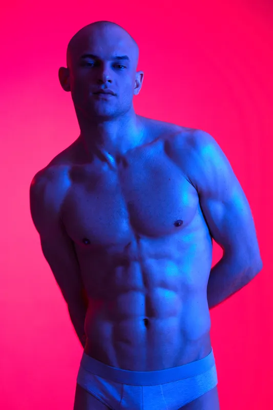 Athletic man posing confidently against a vibrant background, showcasing strong physique and engaging presence. The image is rich in color and energy.