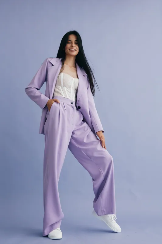 Woman in lilac suit confidently posing against soft purple background, wearing white sneakers and lace top, showcasing modern fashion and casual style.