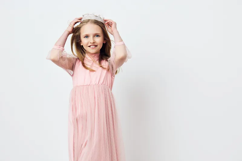Cheerful young girl in a pink dress and crown, smiling against a neutral background, perfect for celebrating childhood moments and special occasions