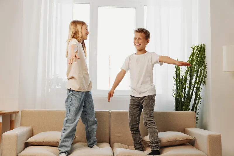 Happy children jumping on sofa in bright living room, having fun and smiling. Cozy home interior with natural light and green plant. Concept of childhood joy and playful moments indoors.