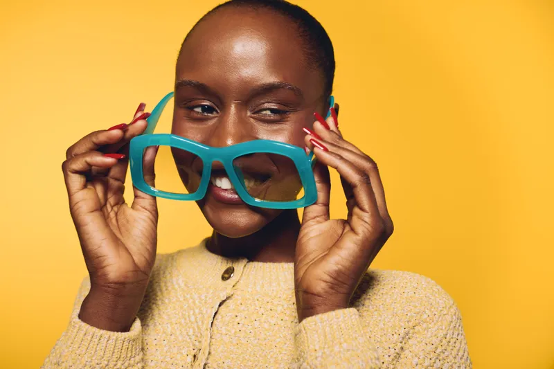 Smiling dark skinned African woman holds oversized blue sunglasses near her face. Yellow background contrasts with beige sweater. Concept of fashion, beauty, and joyful mood.