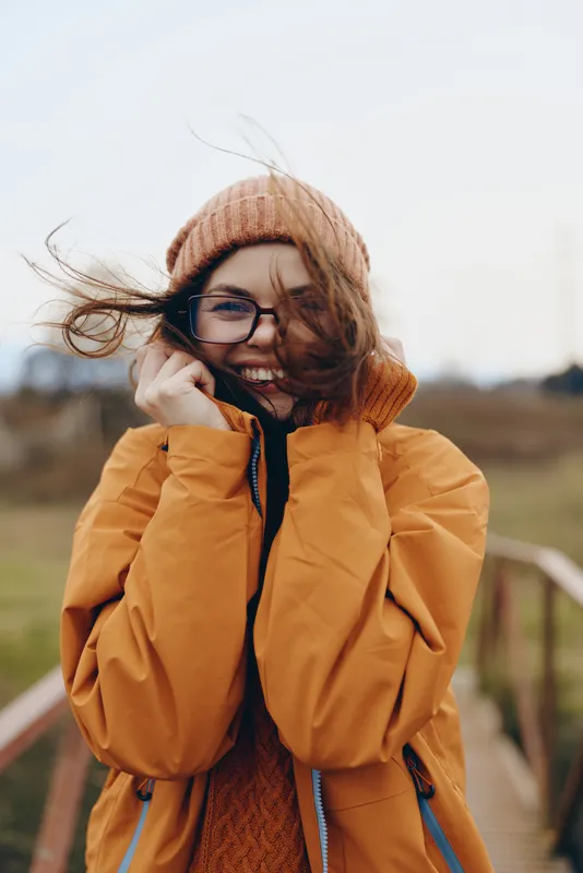 Smiling woman outdoors in winter lifestyle, wearing orange puffer coat, knit hat and glasses, hair lifted by wind, standing near wooden fence, sunlit open field background, natural tones
