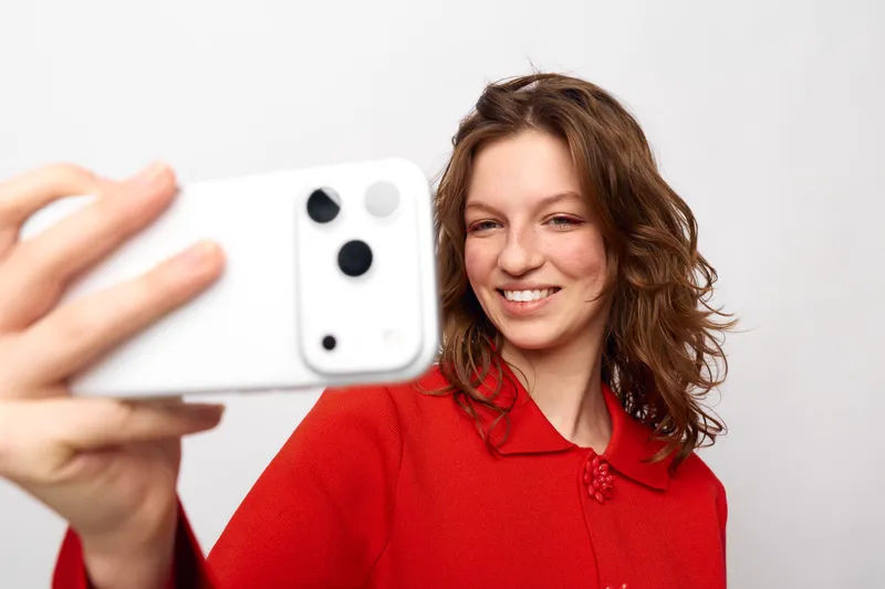 Young woman with curly hair wearing red sweater smiles while taking selfie with white smartphone on white background. Concept of social media, communication, and casual lifestyle.