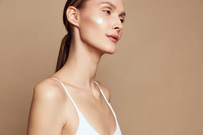Young woman with clear skin applying white face cream on cheek, wearing white tank top, posing on beige background, skincare and beauty concept with soft natural lighting.