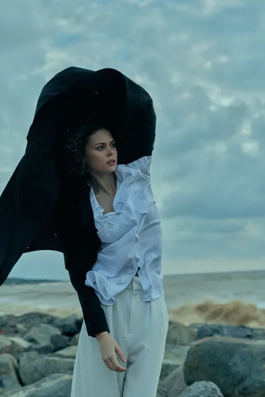 Wind swept fashion on a rocky coast as a woman in white blouse and flowing skirt stands with a dark scarf billowing against a moody sky.