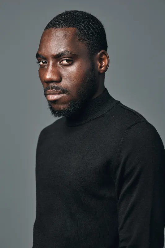 African man portrait in creative conceptual visual style wearing black turtleneck sweater with serious expression on gray background. Studio lighting highlights skin texture and facial features.