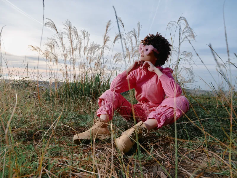 Woman in a pink outfit sits barefoot among tall grasses by a coastal dune, wearing a flower headband and gazing upward, capturing a tranquil outdoor moment with soft light and open sky.