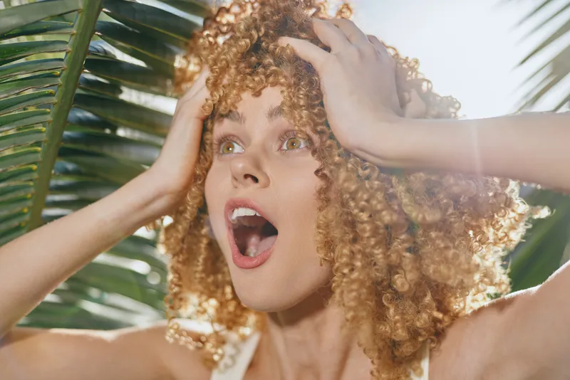 surprised woman with curly hair in tropical sunlight, hands on head, green palm leaves background, natural light, expressive emotions, summer vibe