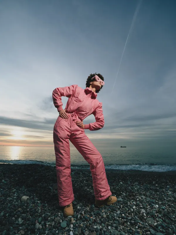 Beach, person, jumpsuit, outdoors, sunset, ocean, fashion, portrait, casual, rocky individual stands confidently on a pebble shore as waves roll in under a vast sky, conveying bold style,