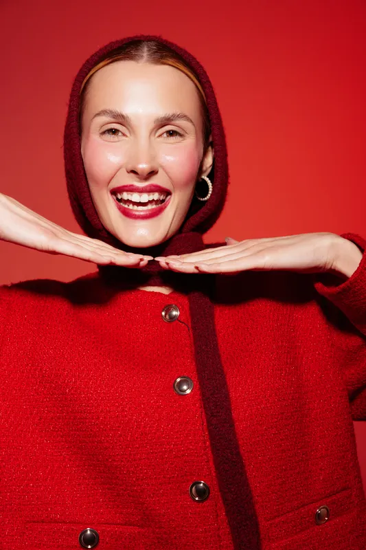 Smiling woman in red sweater poses with hands under chin against red background. Bright studio lighting highlights joyful expression and cozy winter fashion concept.