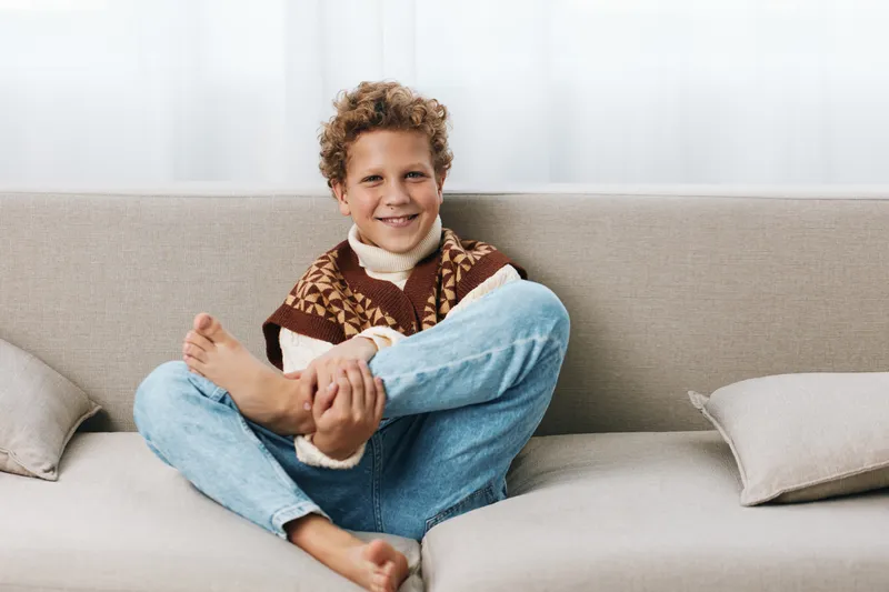A joyful boy sits cross legged on a light couch, smiling warmly. He wears a knit vest and jeans, creating a casual, cozy home portrait with a bright, welcoming mood.