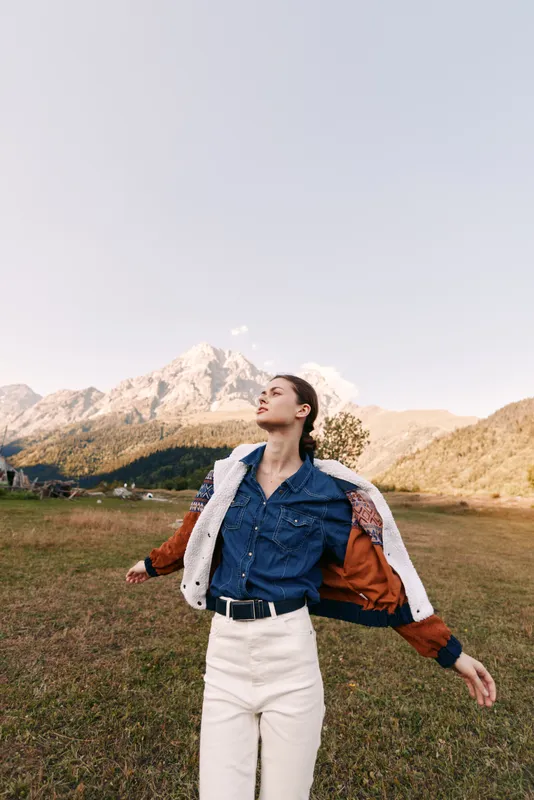 Woman in denim shirt and jacket stands in a mountain field enjoying freedom and fresh air, portrait of a young model outdoors with scenic alpine peaks, grassland and relaxed pose.