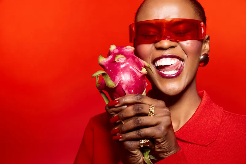 Happy African woman with dark skin wearing red sunglasses and clothing holds a dragon fruit, smiling with tongue out. Bright red background creates vibrant, joyful, and colorful concept.