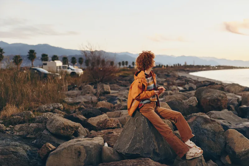 Woman in orange jacket sitting on rocks at sunset, curly hair relaxed, outdoor travel lifestyle, mountains and nature along the coastline, warm evening light, calm mood