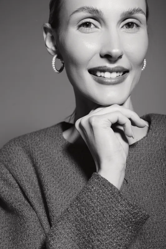 Smiling Caucasian woman with fair skin poses in cozy sweater, hand near chin. Studio portrait in black and white, concept for beauty, happiness, and confidence.