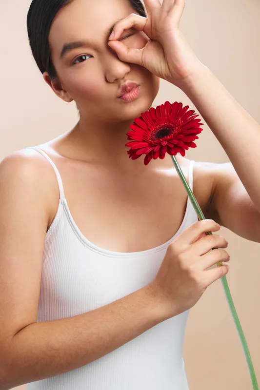 Asian woman wearing a white tank top poses with a bright red flower, making a playful eye gesture and kissy lips, creating a fresh, cheerful mood.