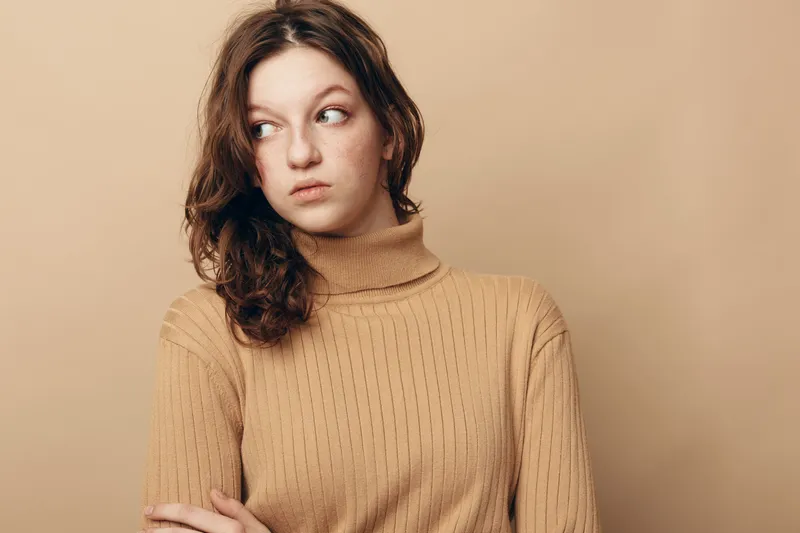 Young woman in beige sweater looks away with crossed arms on beige background. Concept of contemplation, doubt, and casual fashion portrait with natural light and neutral tones.