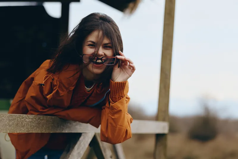 Woman lifestyle outdoor portrait, smiling in orange coat, holding glasses near face, leaning on wooden fence, calm mood, soft daylight, winter background