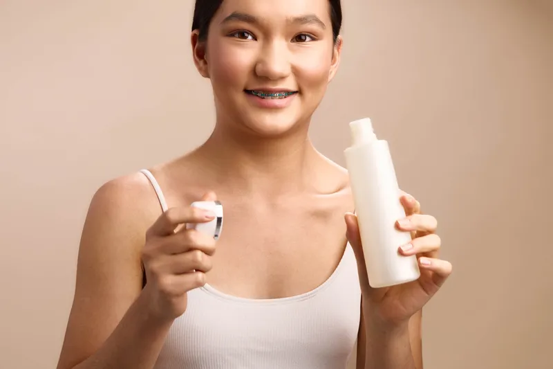 Asian woman poses with skincare products in a clean studio, holding a bottle and jar. She smiles softly, conveying a calm, confident beauty moment and a focus on self care.
