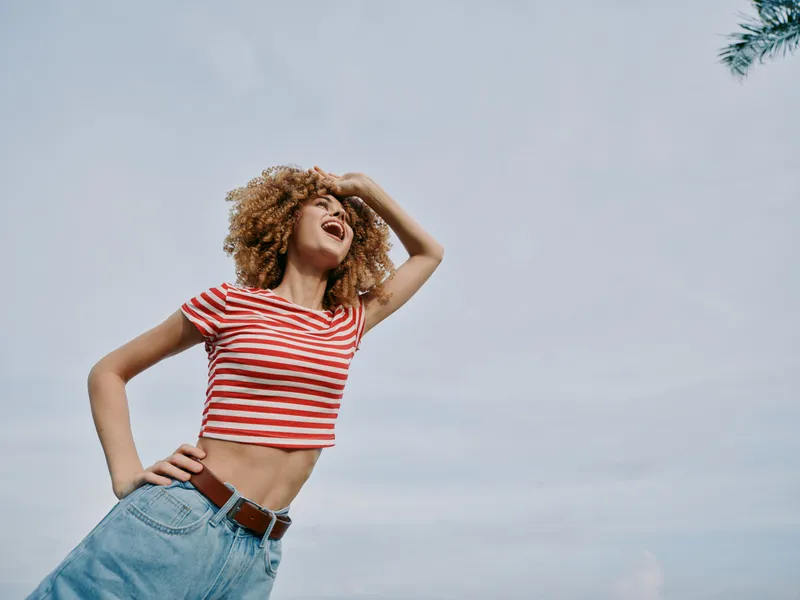 Young woman with curly hair wearing a red striped crop top and high waist jeans poses confidently outdoors under a bright sky, radiating carefree summer energy.