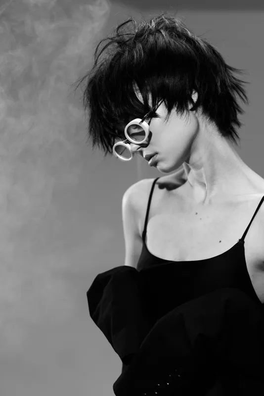 Black and white portrait of a woman with short hair and round glasses wearing a black top in moody lighting.