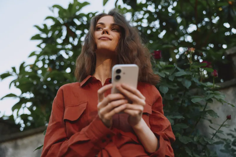 A woman in a red shirt looks up from her phone in a sunny outdoor setting, capturing a candid lifestyle moment for everyday wellness, self expression, connectivity, and personal balance