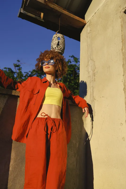 Young woman in bold red jumpsuit stands against a sunlit urban wall, exuding confident energy with a bright yellow crop top and curly hair