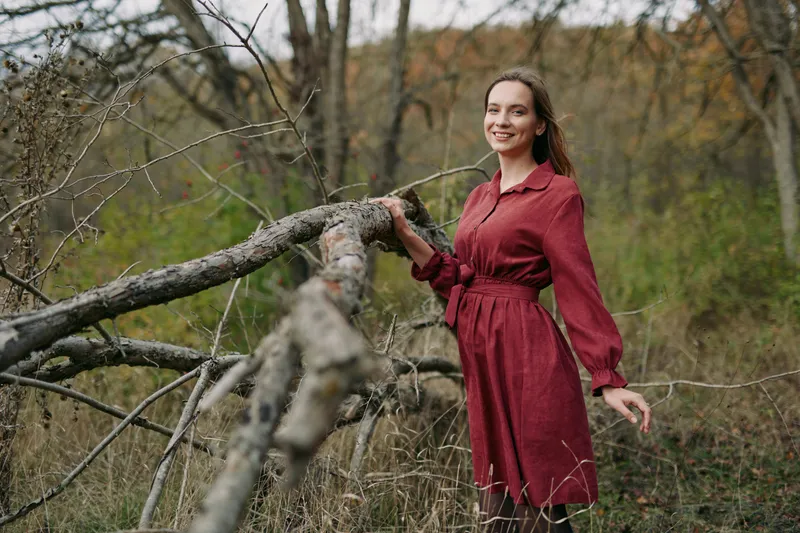 A smiling woman in a red dress touches a fallen tree in a meadow, a candid, genuine portrait that emphasizes naturalness and believable human connection with the landscape.
