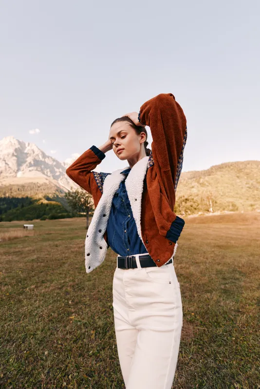 woman portrait outdoor mountains meadow, fashion model wearing brown shearling jacket, denim shirt and white pants, standing pose with hands behind head in warm autumn light