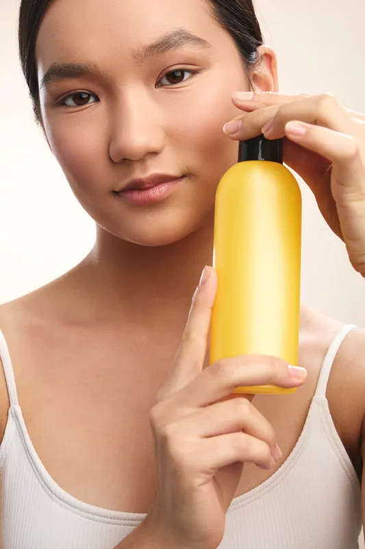 asian woman holds a yellow bottle close to her face in a clean studio, showcasing skincare routine. She looks calm and focused, creating a clean, confident beauty moment.