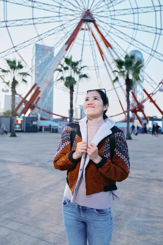 Traveling, sightseeing, outdoor urban plaza scene with a stylish jacket and scarf, a smiling traveler looks up at a towering ferris wheel against a modern skyline