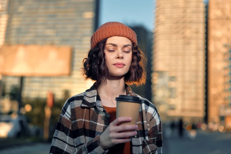 Woman holding coffee in an urban portrait with beanie and sunlight, candid lifestyle and authenticity captured in golden hour glow, mindful living and emotional storytelling moment.