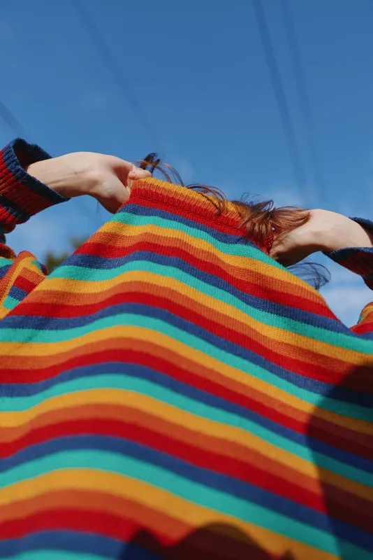 Sweater stripes colorful person pulling knit sweater over head with hands visible, low angle view against blue sky, casual fashion, playful pose and texture details.